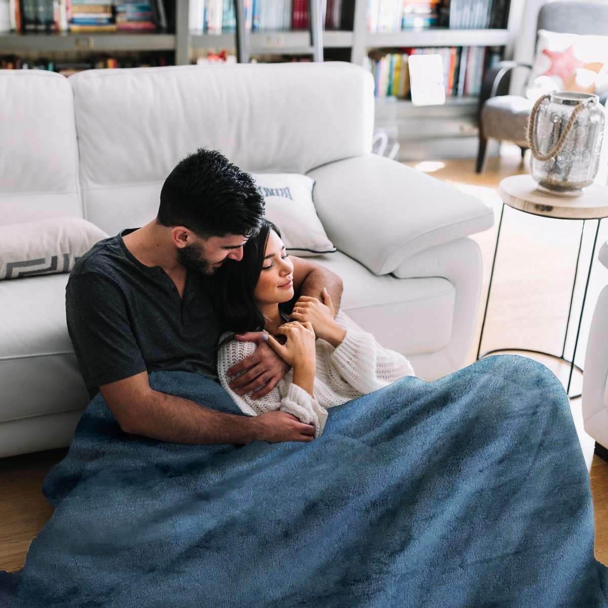 Esposos sentados en el piso, abrazados con cariño y cubiertos por una cobija color petróleo, disfrutando de un momento íntimo y acogedor juntos